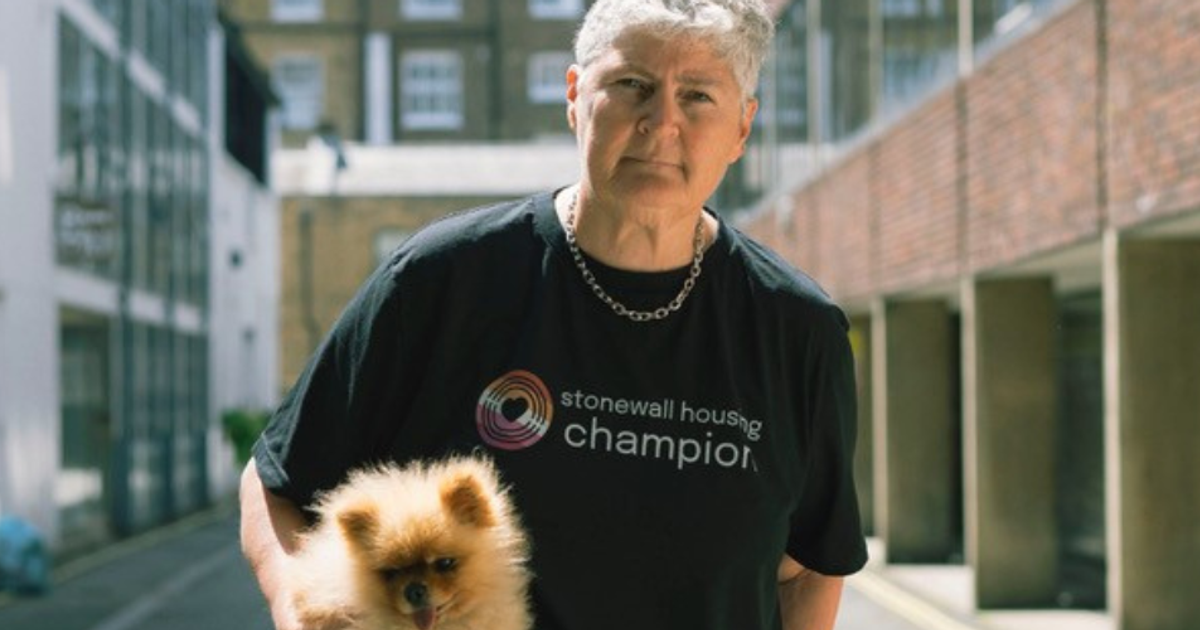 Linda Riley, advocate for Lesbian Visibility Week, wearing a Stonewall Housing shirt and holding a small dog in an urban alleyway.